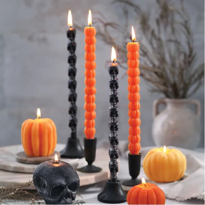 Big Dipper Wax Works Sculpted Orange Pumpkin Taper Candles lit and displayed in black candlestick holders on a Halloween tablescape, with two tall orange pumpkin-silhouette tapers burning alongside decorative pumpkin-shaped candles in orange and yellow, dried branches in a ceramic vase, and a black skull prop, against a moody grey wall background